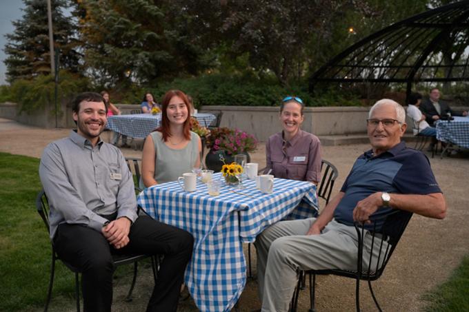 Group of four people sitting at blue checkered tablecloth outdoors.