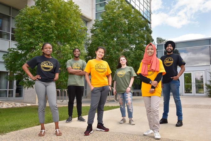 Six students standing outside with glass building in the background.