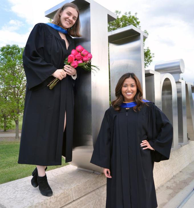 Two students in full convocation gowns stand next to sign.