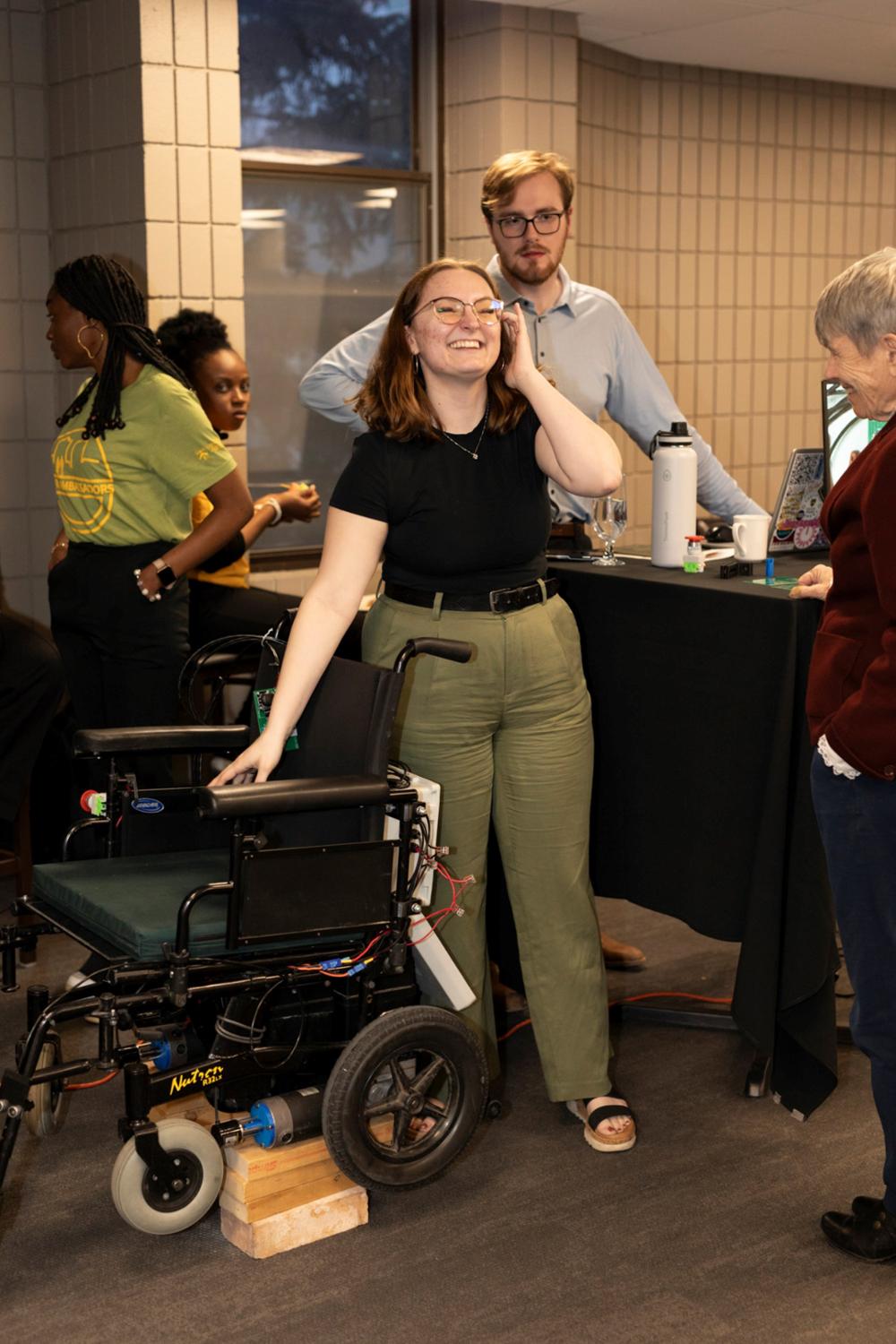 Two smiling individuals stand by automated wheelchair at event.