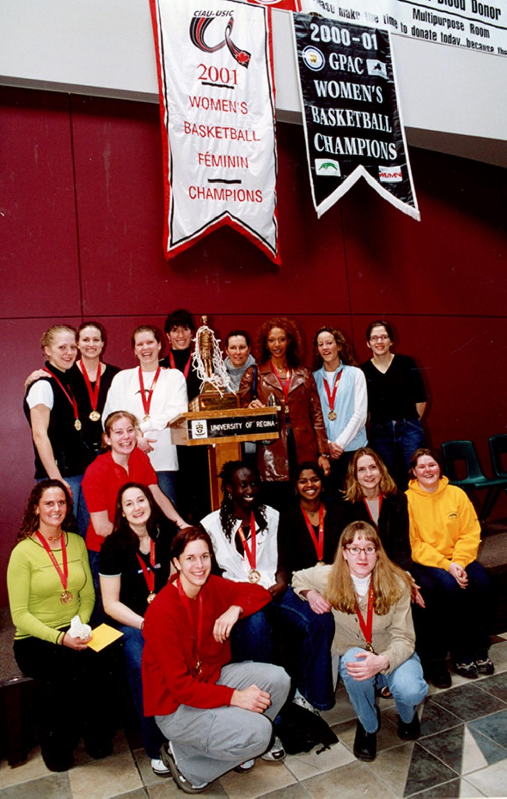 Women’s basketball team posing with trophy, medals, and winning banners.