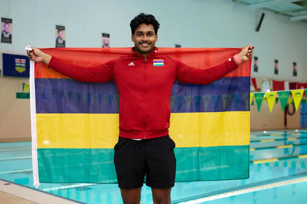 A young man poses on a pool deck with the flag of Mauritius draped across his back.