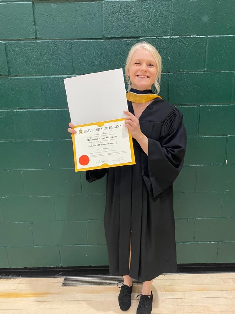 A person standing against a wall wearing a graduation gown and holding a Bachelor of Science in Nursing degree.