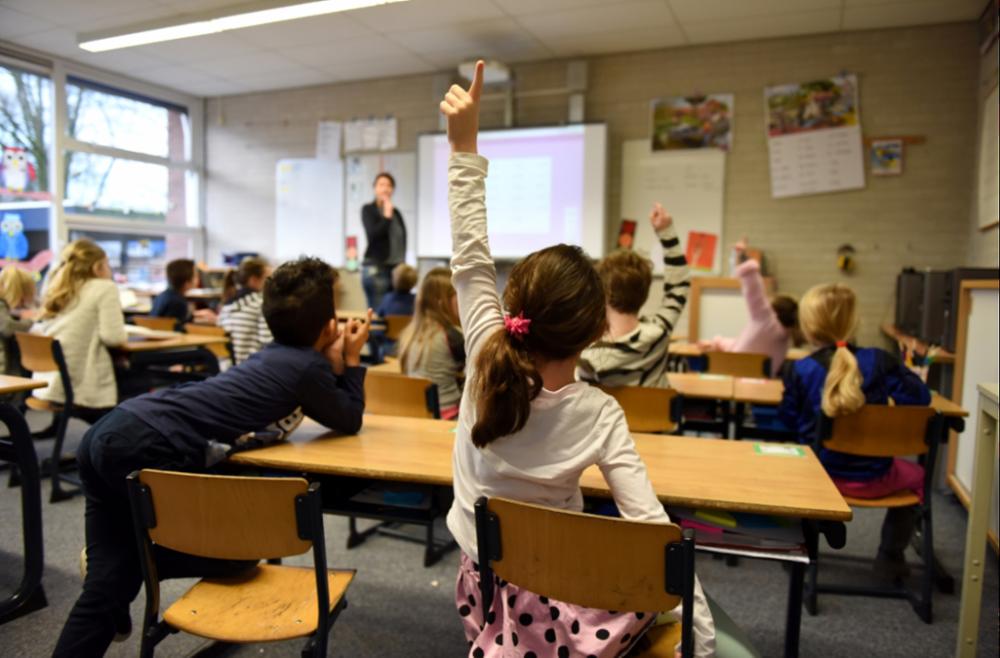 Children sitting at desks looking at a female teacher standing at the front of a classroom.