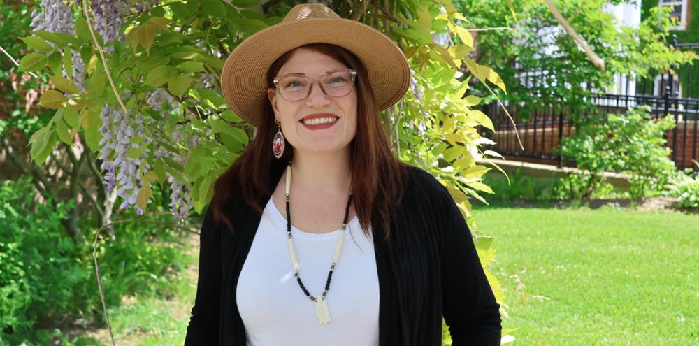 Headshot of smiling woman wearing a hat standing outside with tree and grass in the background.