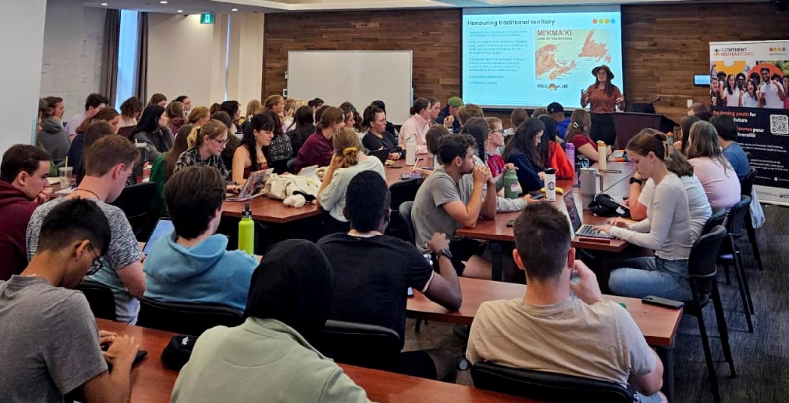 Students seated in rows with female instructor wearing a hat standing at the front of the room and a large screen behind her.