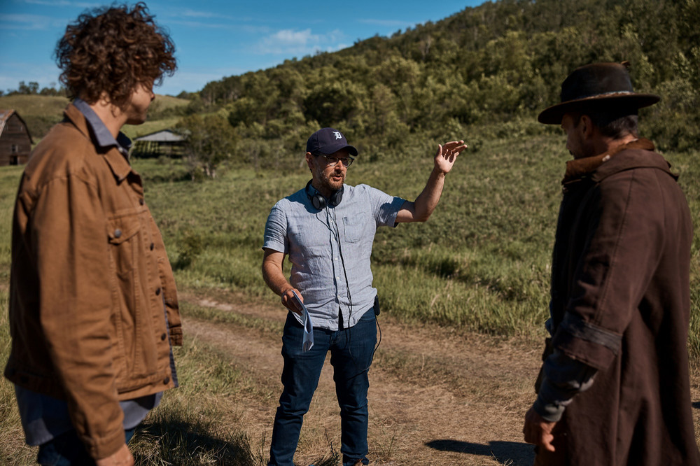 Three men standing in a group with one man explaining something with hand gestures and a tree-covered hill in the background.