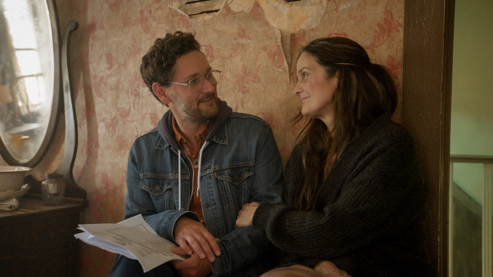 Man with glasses and woman with long hair sit close while leaning against a wall with rippled wallpaper.