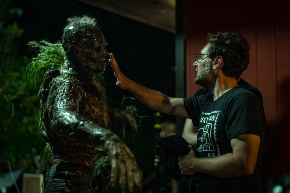 Man with glasses touching the face of a man covered in moss and dark make-up outside at night. 