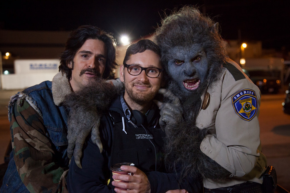 Two men standing facing camera with a third man on the right in wolf make-up dressed in a cop’s uniform. 