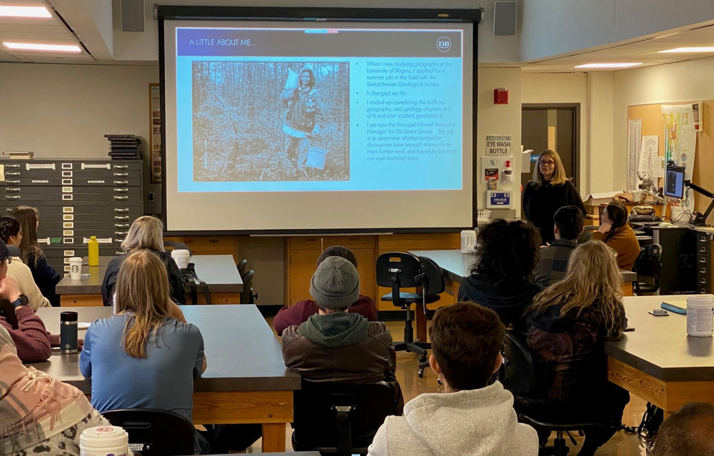 Woman standing in a lab at a screen presenting to young women and men seated at tables.