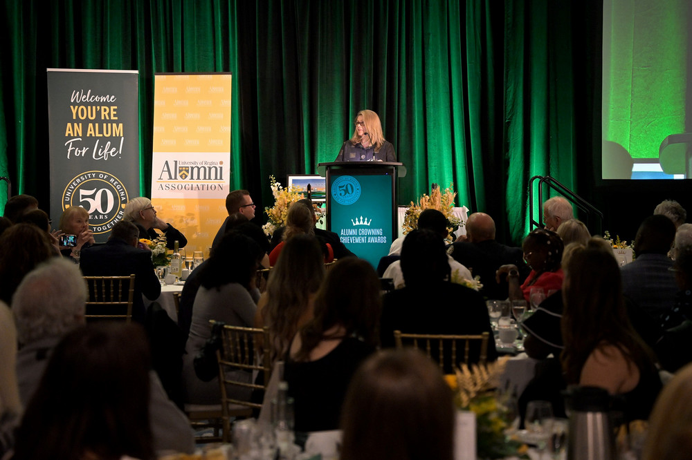 Woman standing at podium presenting to a large group of people seated at tables at an evening  celebration.