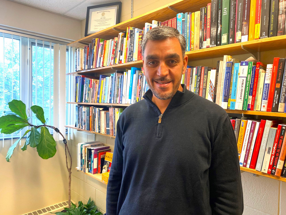 Man smiling seen from the shoulders up with bookshelves, a window, and plant in the background.