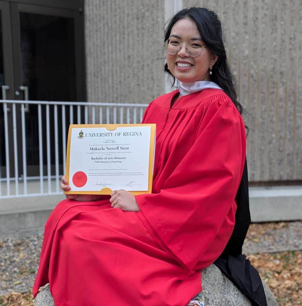 Young woman with red robe sitting on rock and smiling while holding a certificate in her hands.