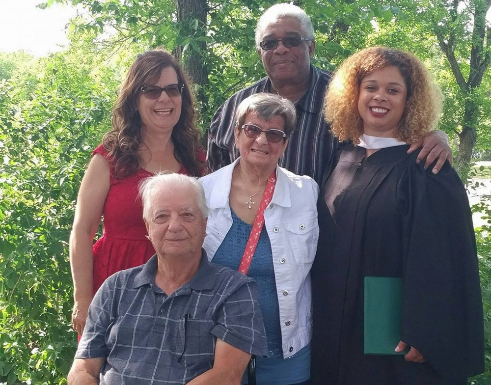 One man sitting and three women and one man standing as a group outside with trees in background and the woman standing on the right wearing a graduation gown.