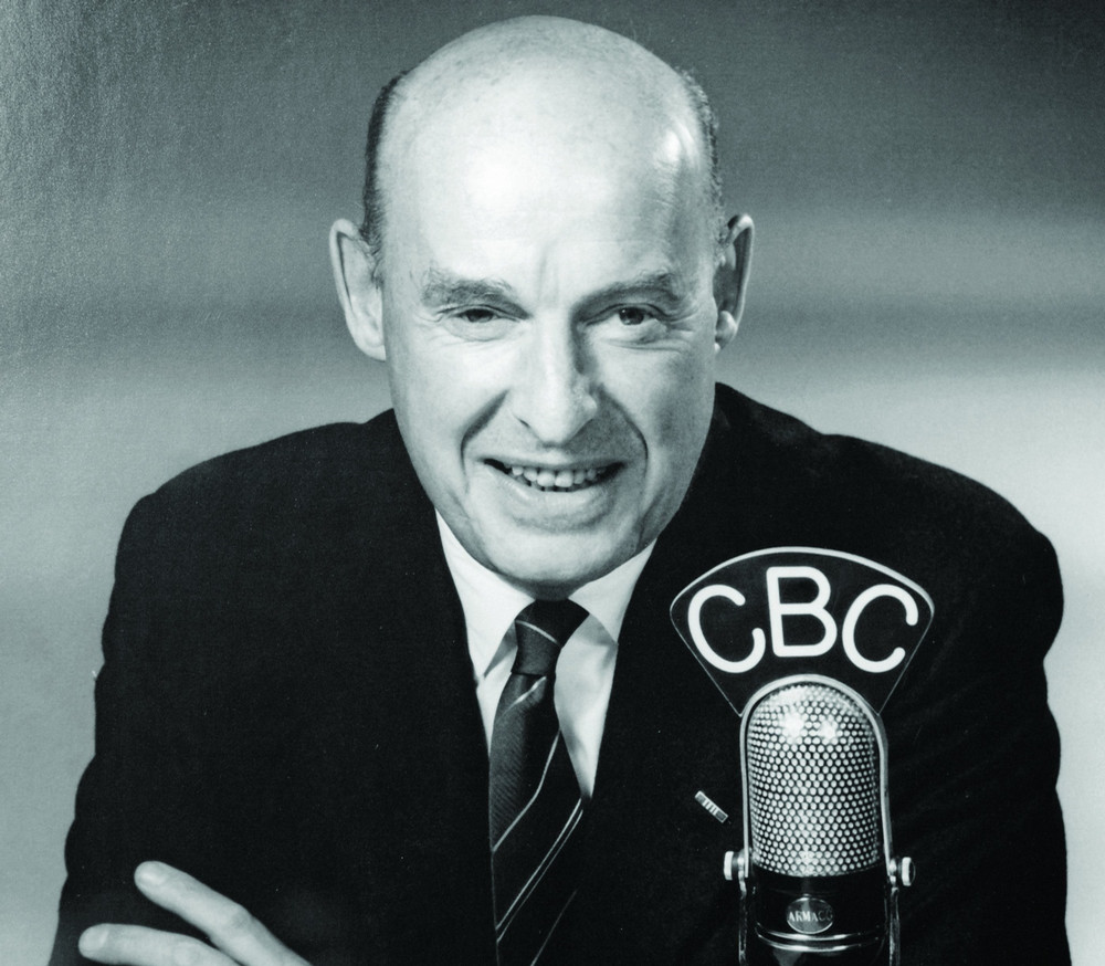 Headshot of middle-aged man from the 1960s seated and wearing a suit while talking into a microphone.