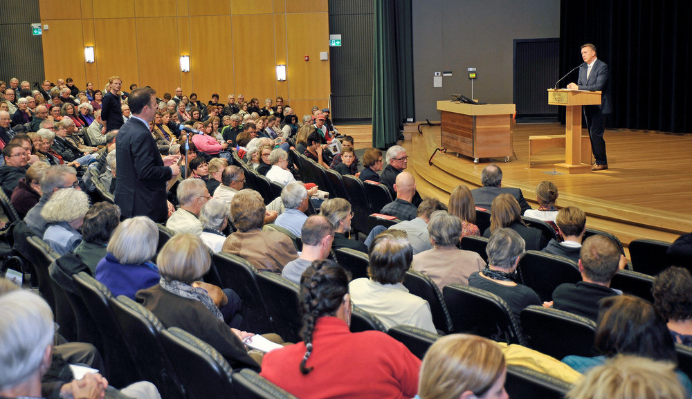 Crowd seated in brightly lit auditorium with a man in a suit standing at a podium on a stage.