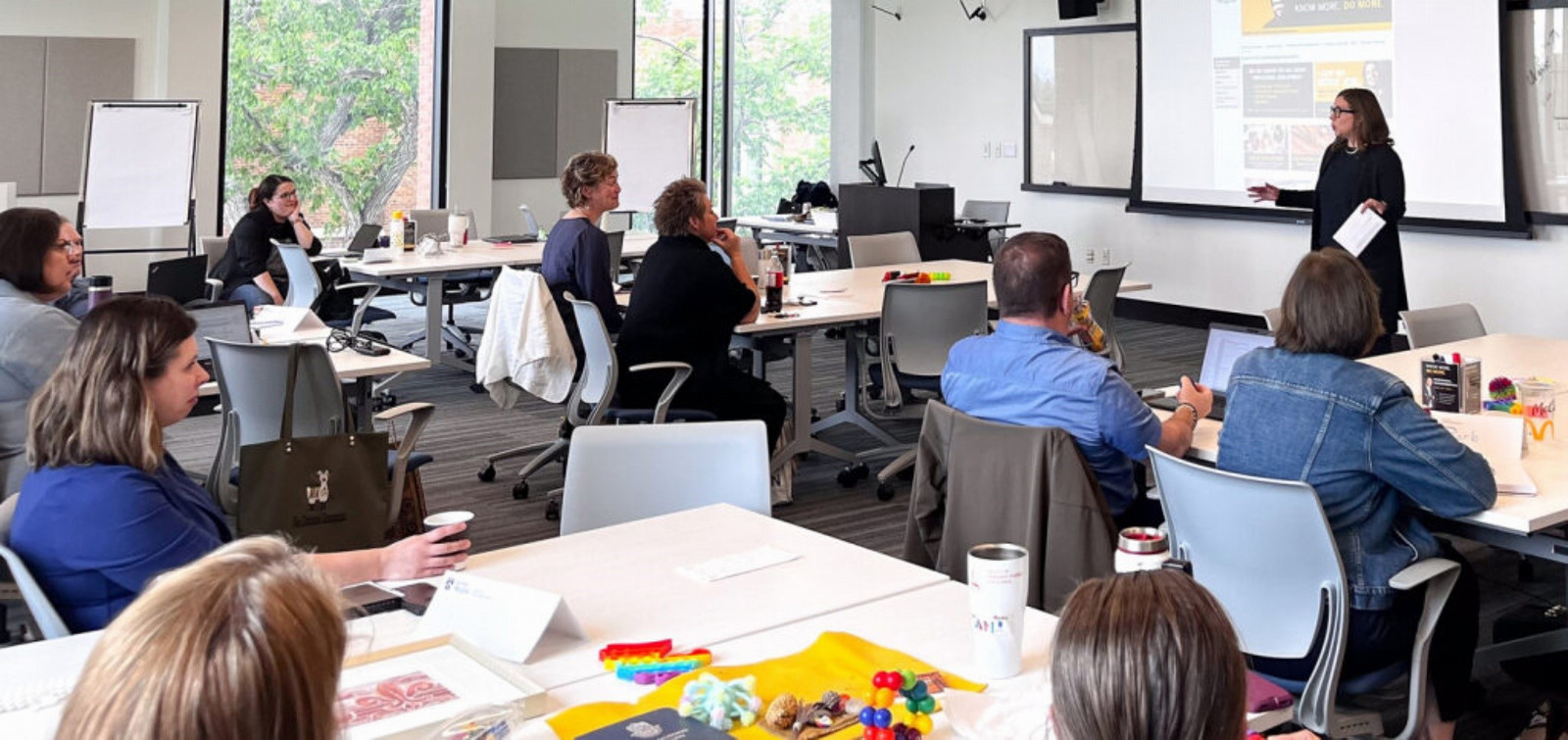 Adults sitting around desks in classroom with a female instructor at the front of the room standing in front of a screen.