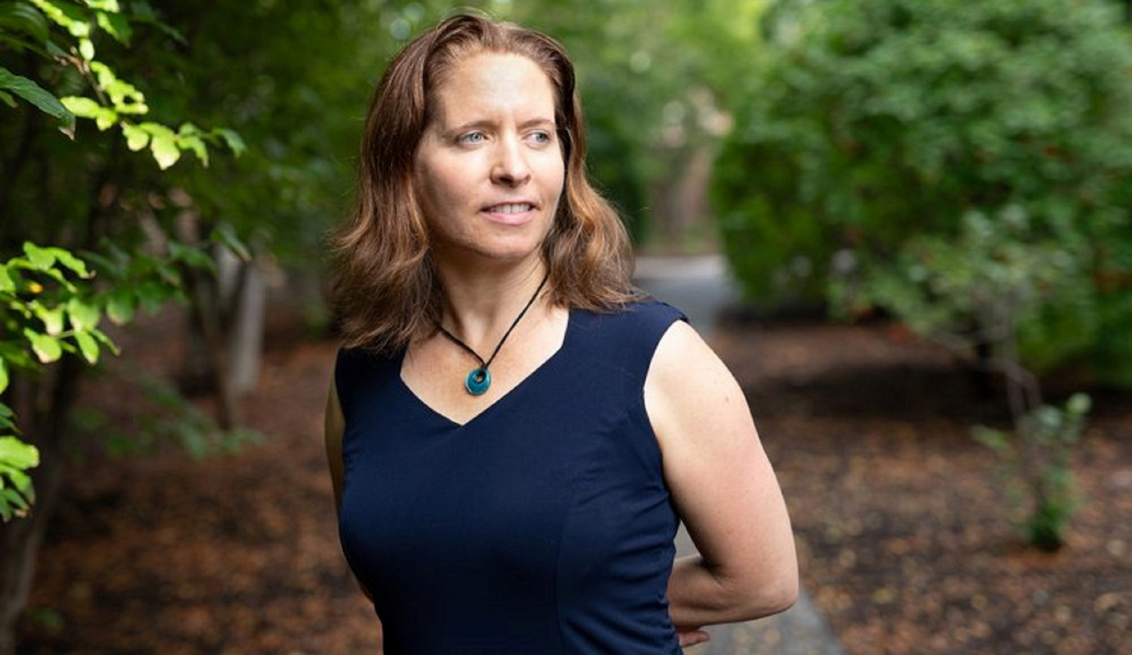 Woman with hair to shoulders standing on a path in the forest and looking to the side while smiling.