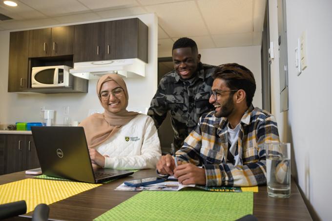 Three students looking at computer in kitchen.